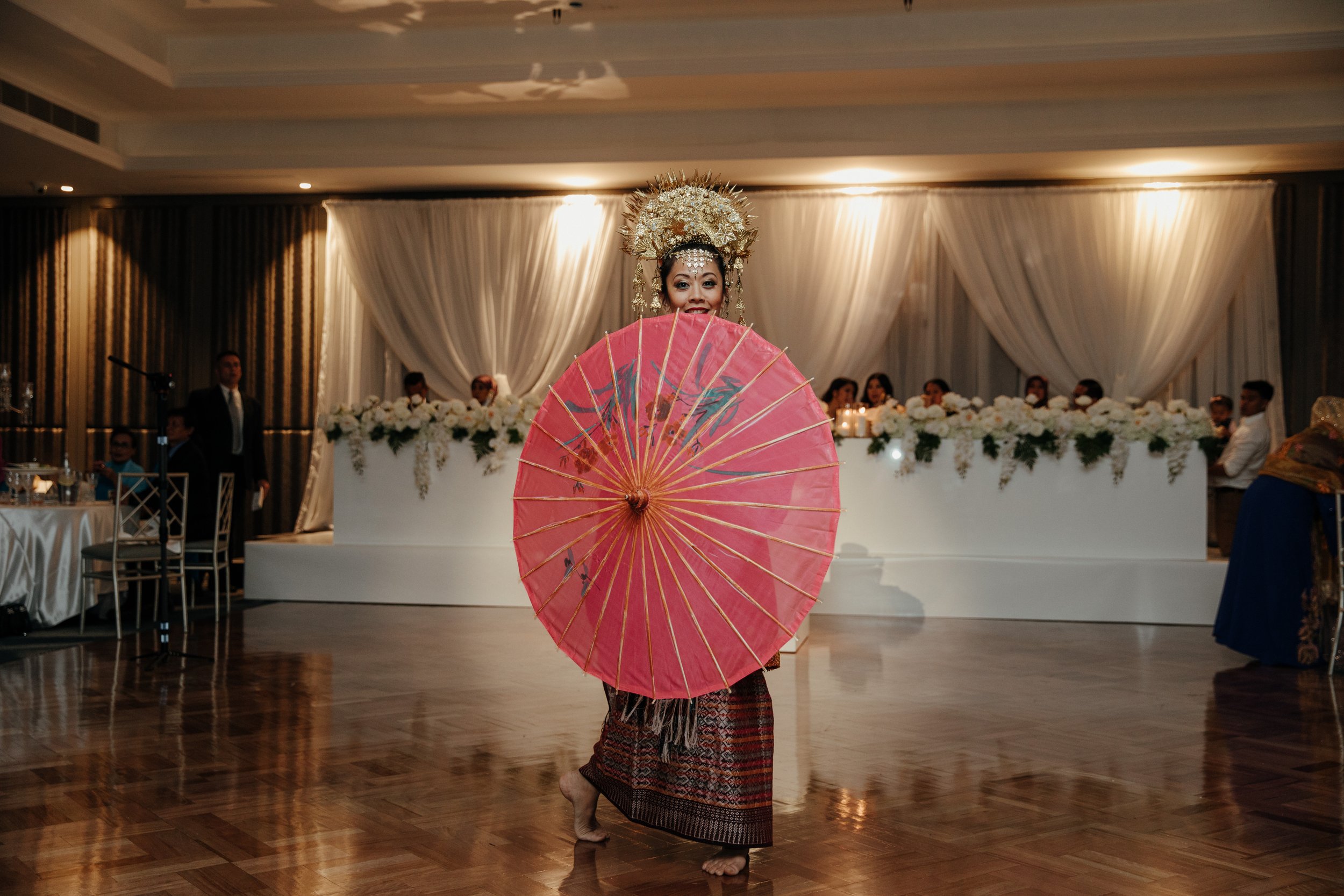 Group of six women and one man in traditional Indonesian attire, posing indoors. The women wear red and gold patterned clothes with floral accessories and hold scarves, while the man wears a black and gold traditional outfit with a headpiece. 
