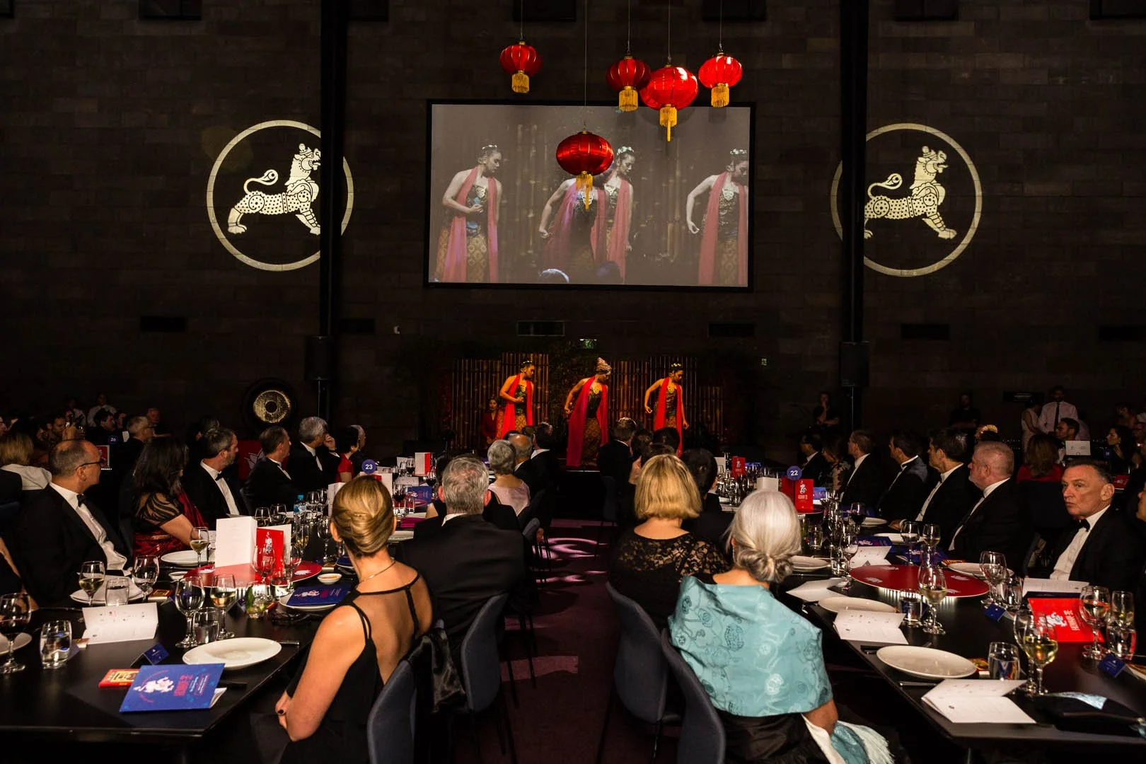 Guests seated at round tables watching a stage performance with three dancers dressed in red and black costumes, hanging red lanterns, and a large screen displaying the performers.