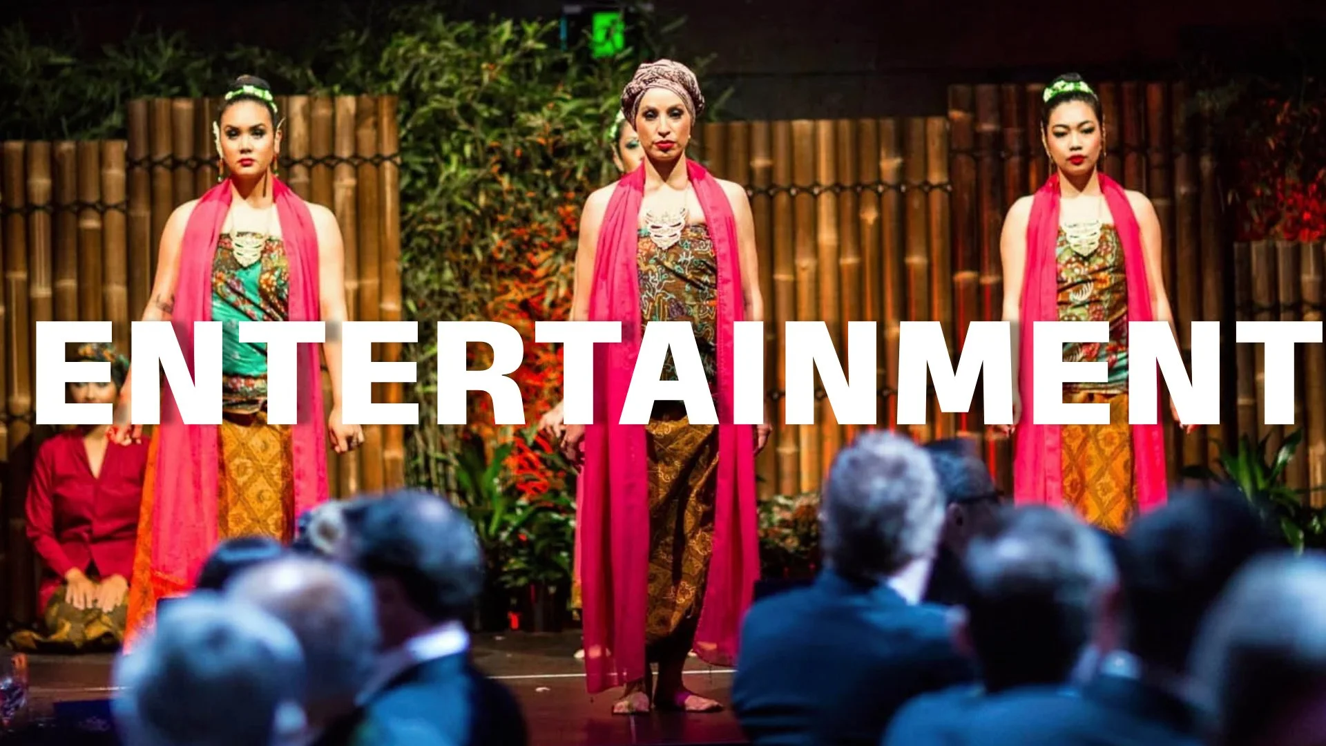 Three women wearing traditional Indonesian batik costumes and pink scarves on stage, with an audience in the foreground and a bamboo fence in the background.