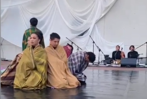 Two women in yellow robes are kneeling on a stage with their eyes closed, possibly praying or meditating. In the background, other people are also seated or standing, with some near microphones and sound equipment.