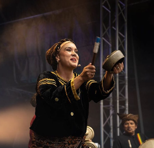A woman in traditional attire playing a percussion instrument on stage at a cultural event, with a man in similar attire in the background.