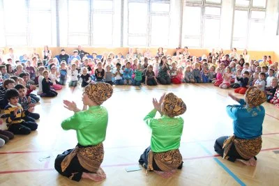 Three women in traditional Indonesian clothing perform a dance in front of a large group of children seated on the floor in a gymnasium.