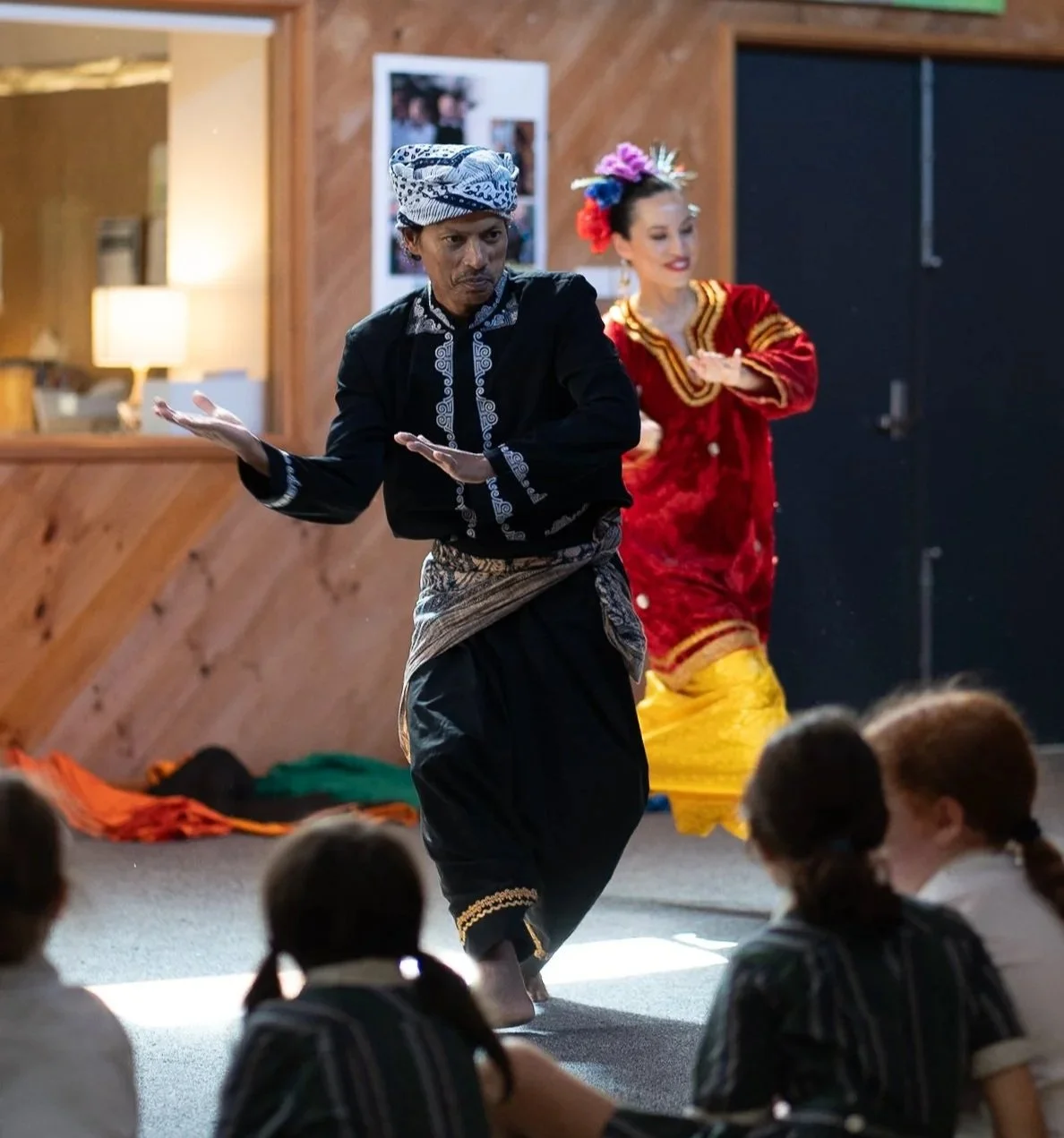 Two performers dance in front of children in traditional Indonesian attire, one in a black and white costume and the other in vibrant red and yellow clothing with floral headpiece.