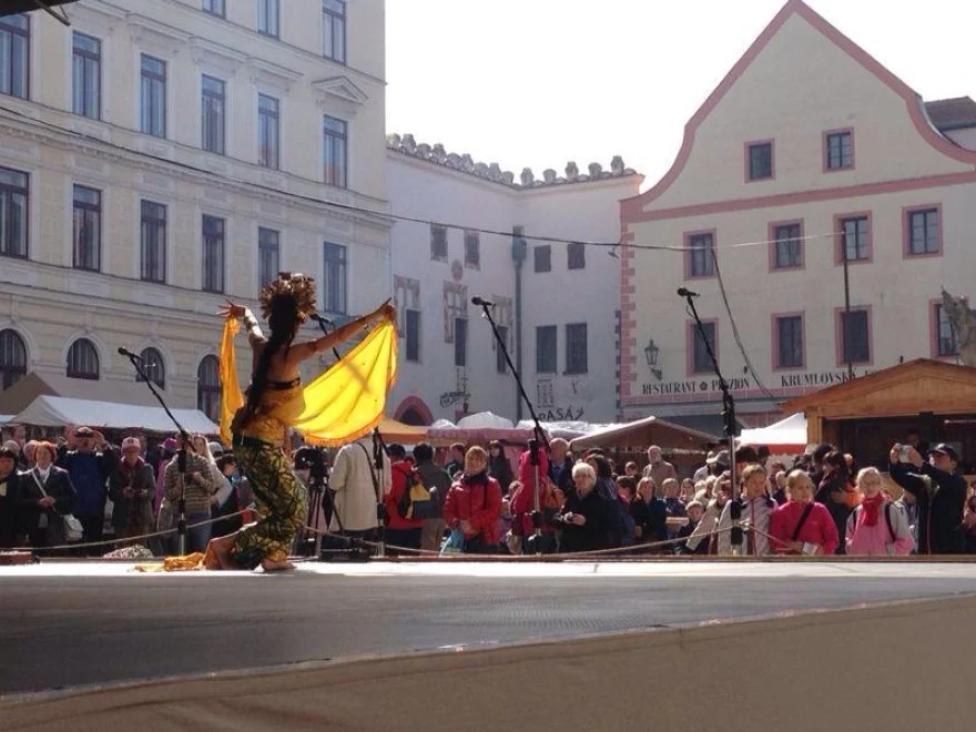 A performer dancing on stage during an outdoor cultural event or festival, with a crowd of spectators and traditional European-style buildings in the background.
