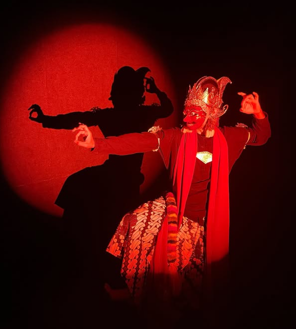 Performer in a traditional Indonesian mask and dance costume on stage with dramatic red lighting and shadow behind them.