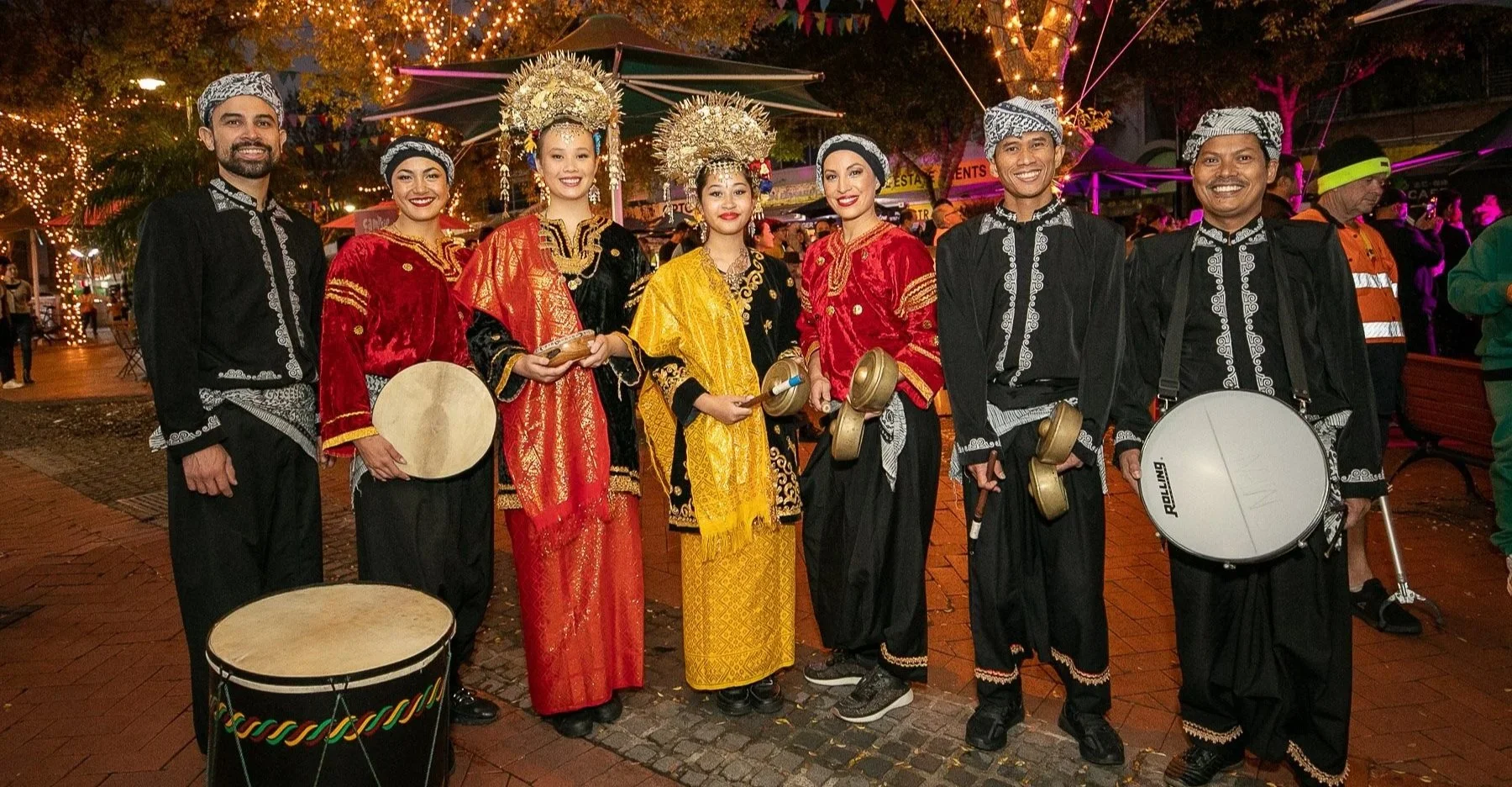 Group of eight people in traditional Indonesian clothing, holding musical instruments, standing outdoors at night with festive lights and decorations in the background.