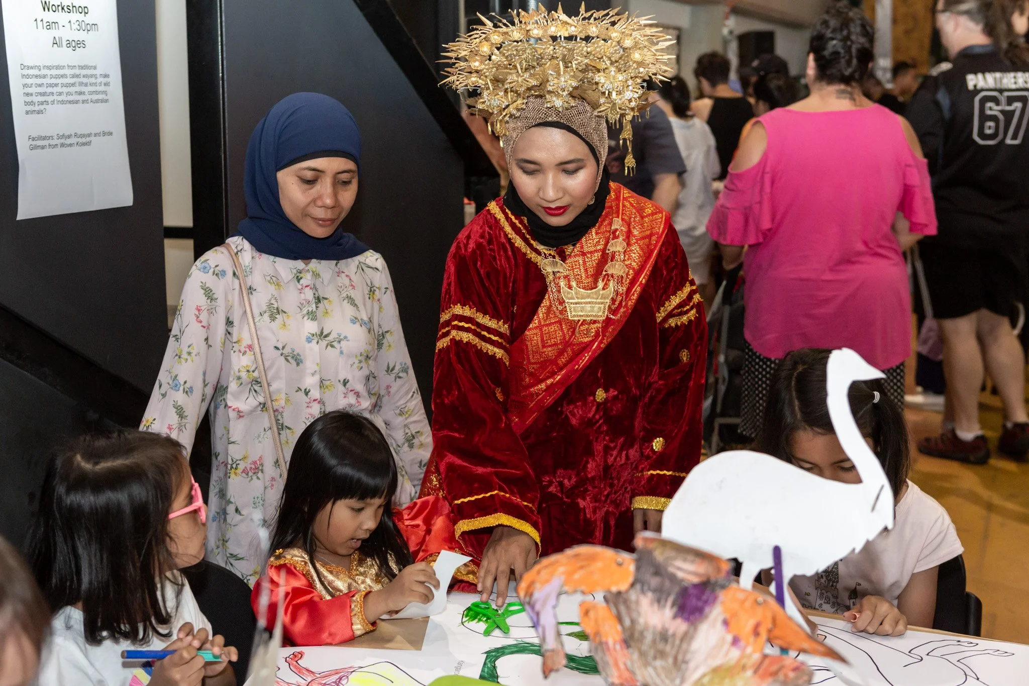 A woman in traditional Indonesian attire, including a red velvet dress and an ornate gold headdress, participates in a children's craft activity at a cultural event, with children and other adults around a table of art supplies and paper crafts.