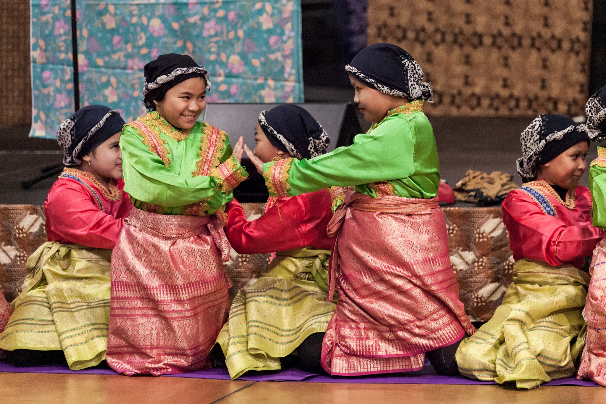 Children dressed in traditional Indonesian costumes, with pink and green outfits, wearing black headscarves, sitting on the floor and dancing