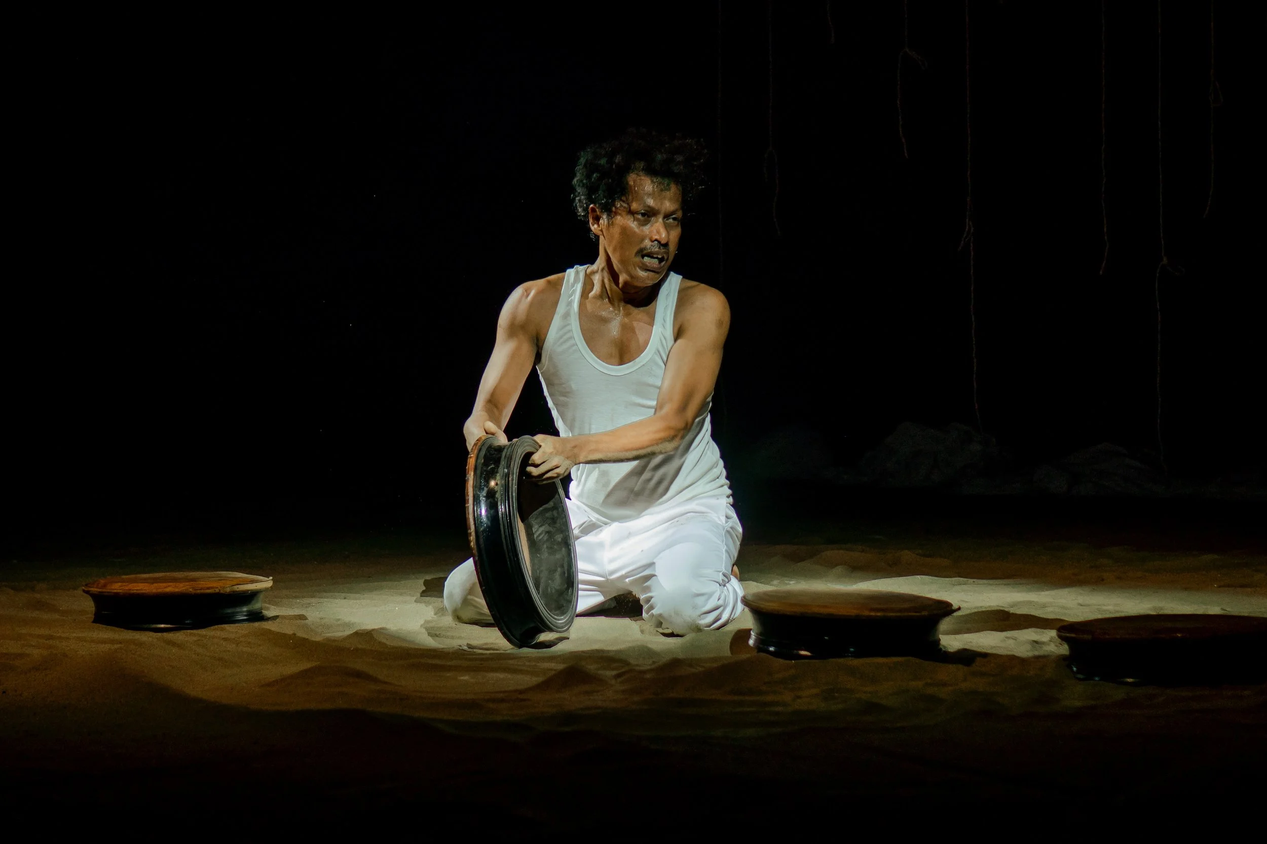 A performer kneeling on stage, holding a large flat drum,  with a focused expression, in a dark setting.