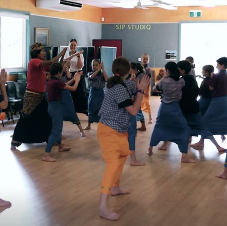 Children and instructor practicing a dance  in a studio with wooden floors and large windows.