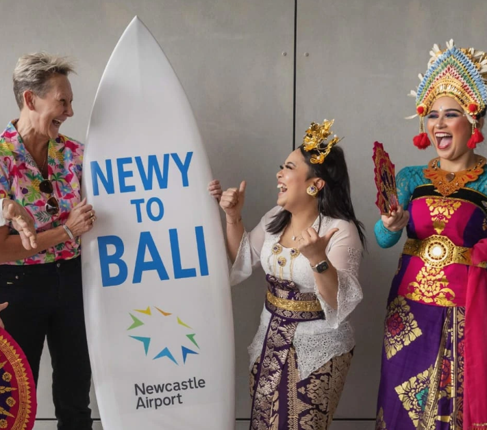 Three women smiling and talking at an airport with a large sign that says 'NEWY TO BALI' and features the Newcastle Airport logo. Two women are dressed in traditional Balinese attire, including colorful headgear and intricate clothing, while the third woman is dressed casually.