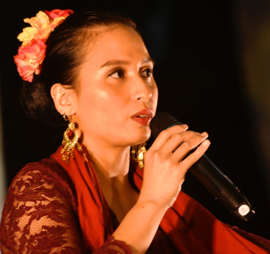 A woman in traditional Indonesian clothing holding a microphone, wearing floral earrings and a flower hair accessory, possibly performing on stage.