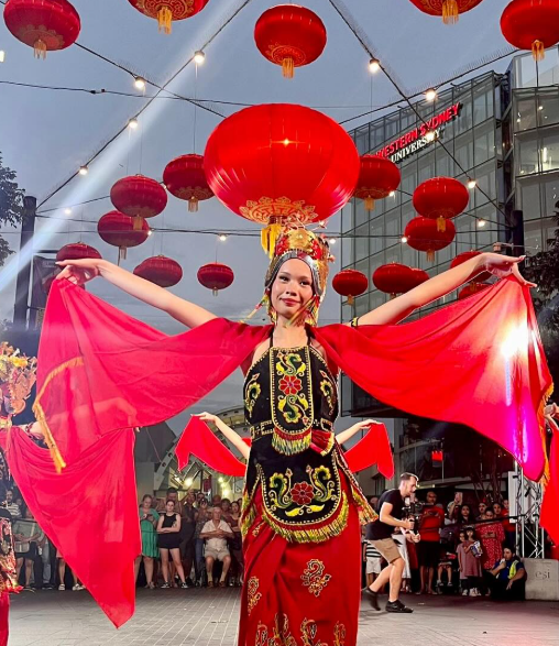 A woman in traditional Chinese dress performing in front of hanging red lanterns and a crowd at an outdoor event.