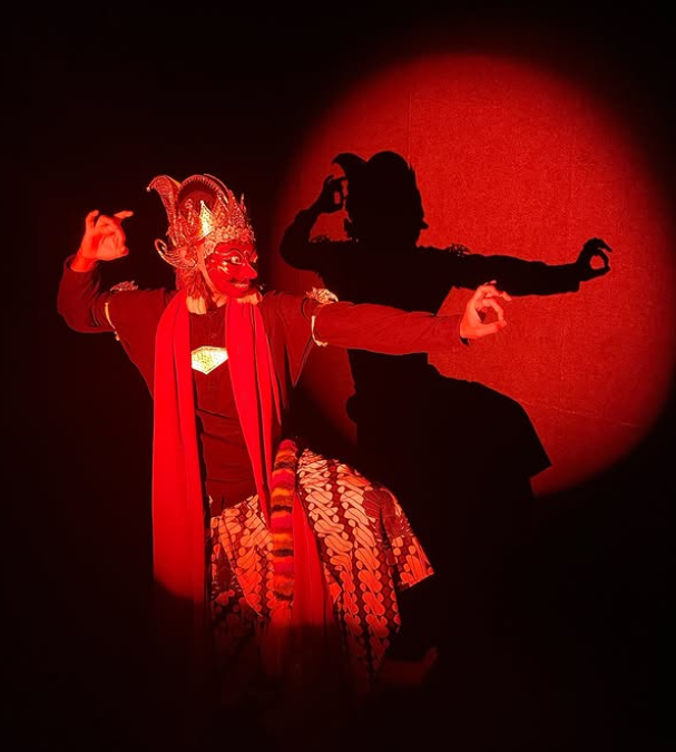 Performer in traditional Indonesian mask and costume, striking a pose with arms extended, shadow cast on red-lit background.