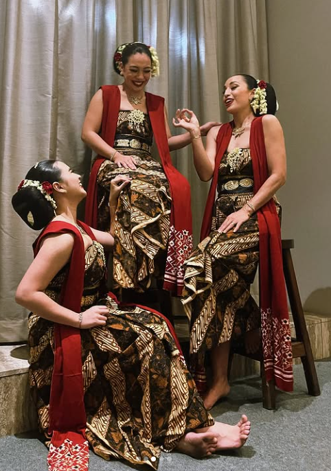 Three women in traditional batik dresses and red vests, with flowers in their hair, practicing a dance on a stage with a curtain backdrop.