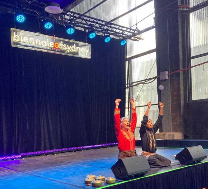 Two performers kneeling on stage with their arms raised in celebration at the Biennale of Sydney, with a black curtain background, stage lights, and a neon sign overhead.