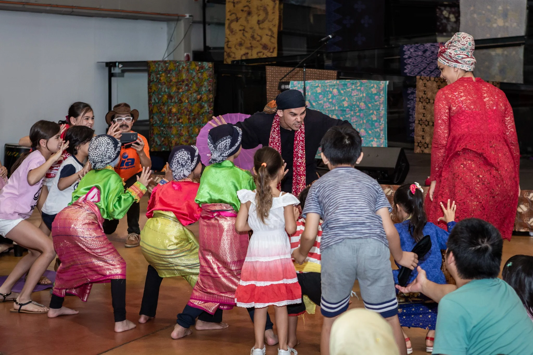 Children dressed in colorful clothing playing with tho performers on stage, with one performer in black and another in red lace.