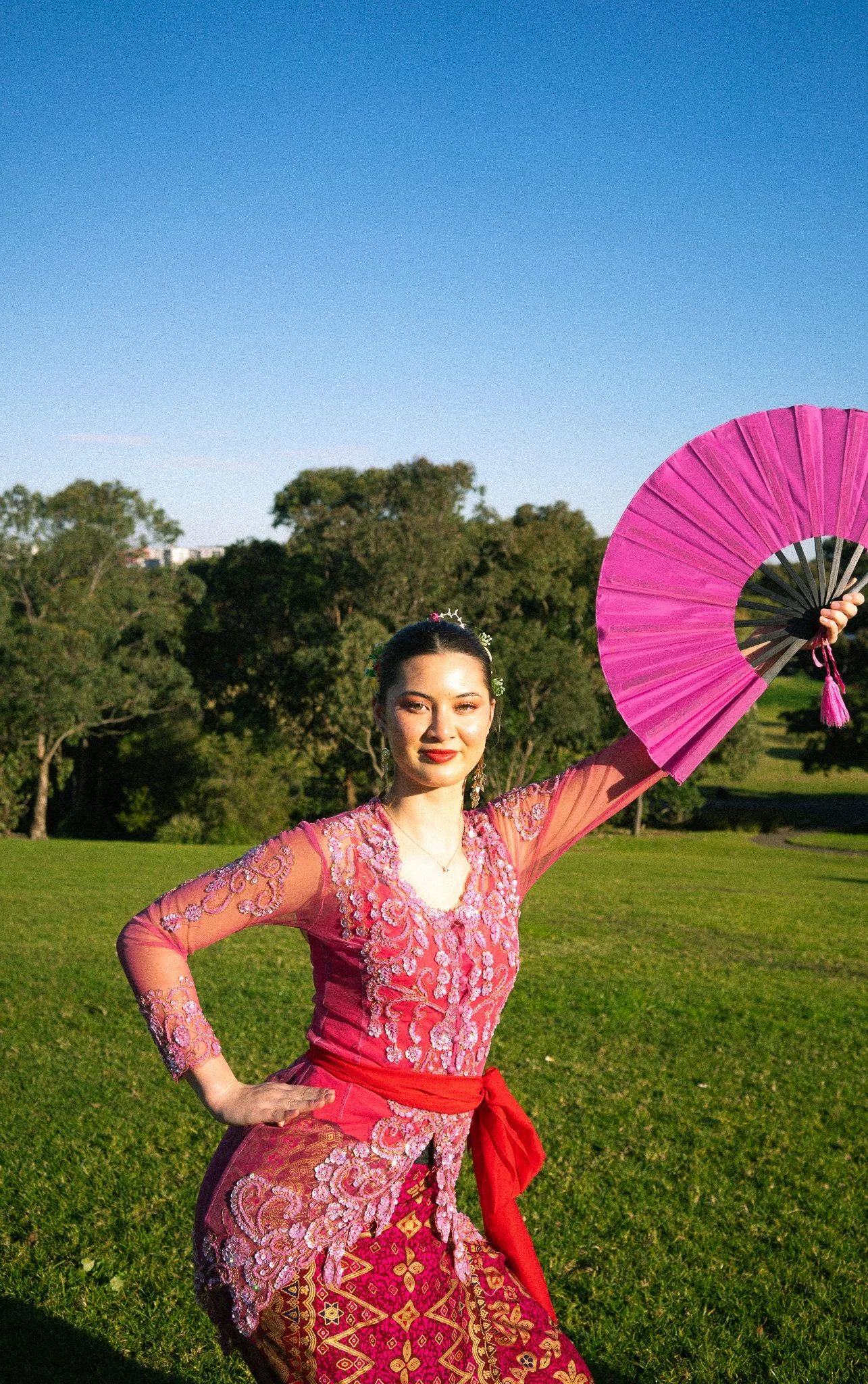 A woman dressed in traditional Indonesian attire holding a pink fan outdoors in a grassy park with trees and a clear blue sky.