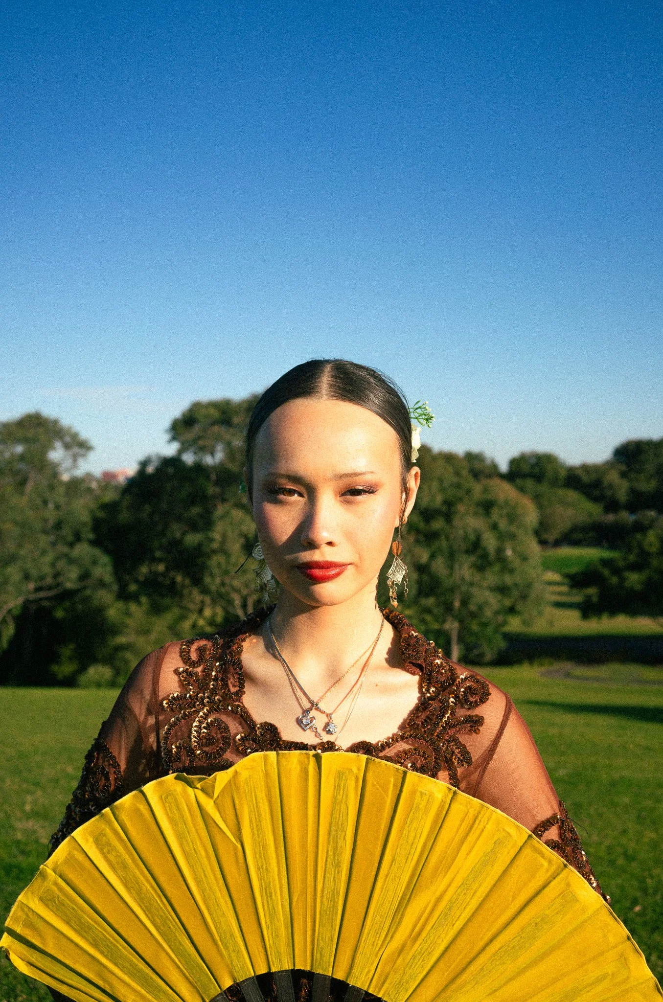 A woman dressed in traditional attire holding a yellow fan, standing outdoors with trees and a clear blue sky in the background.