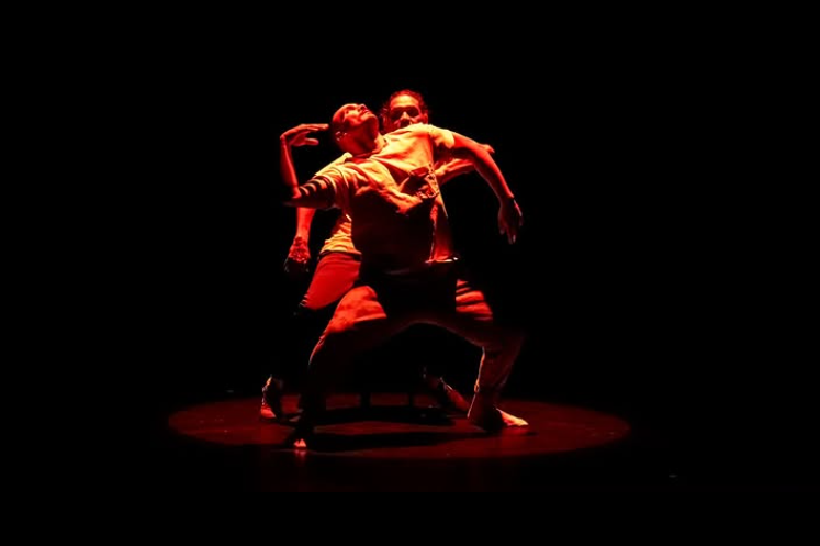 Two dancers perform on a dark stage with red lighting, with one dancer kneeling and the other standing behind them.