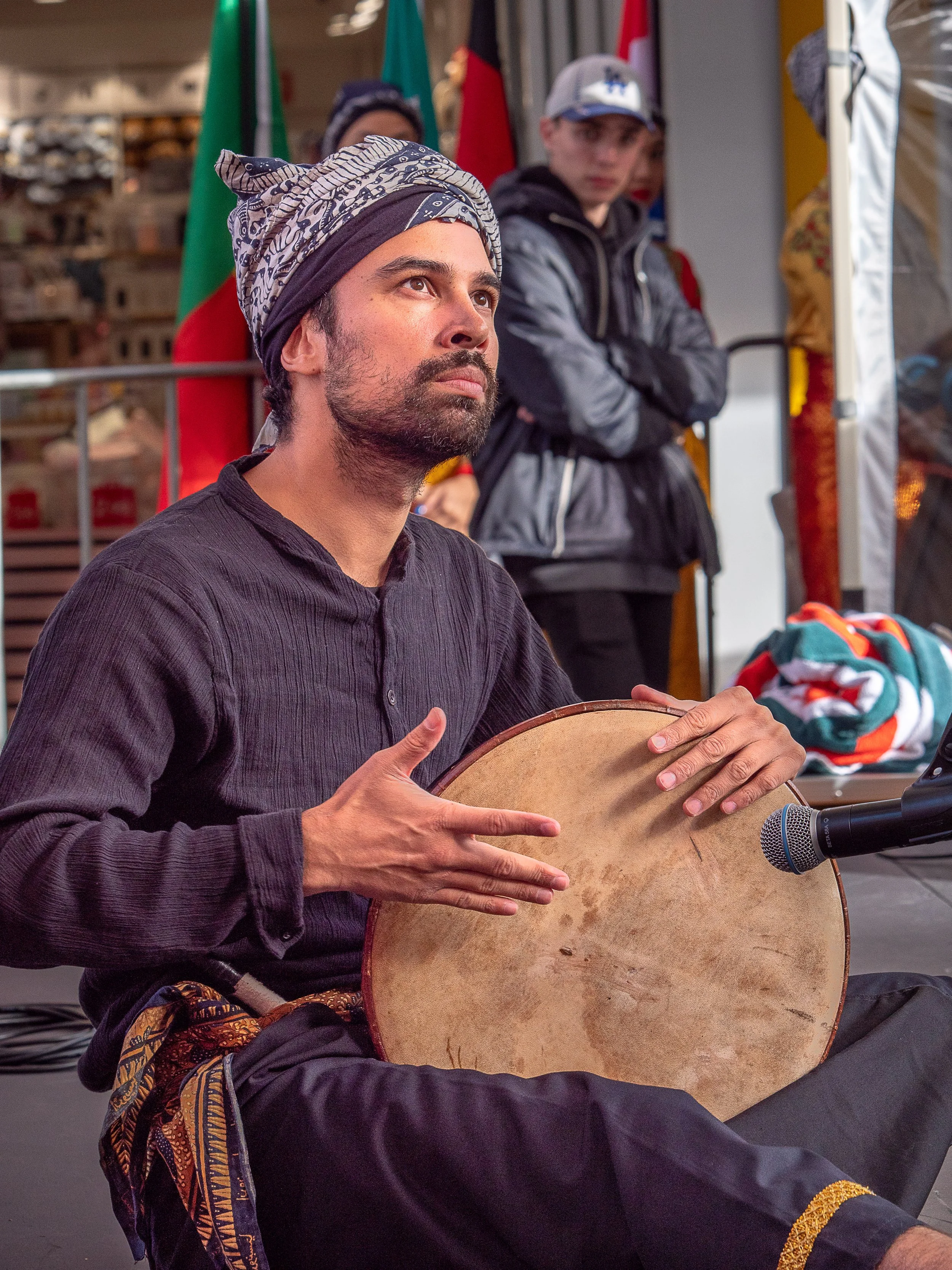 A man playing a hand drum with a microphone in front of him, seated on the ground, with a woman and a boy standing behind him at an outdoor event.