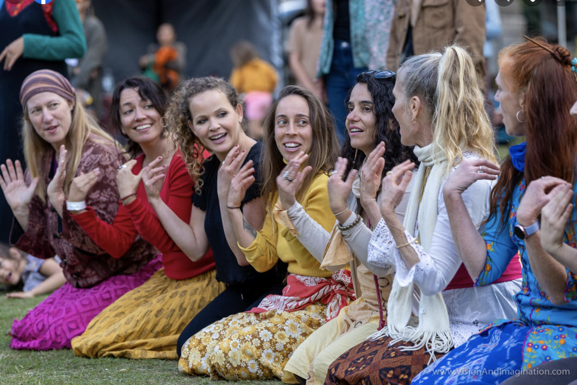 Group of women sitting on grass at outdoor event, clapping and smiling, dressed in colorful clothing.