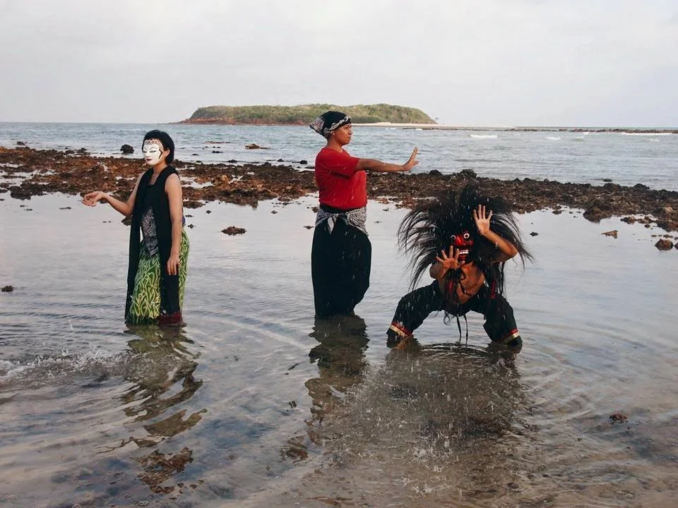 Three performers in costumes and masks on a rocky beach with the ocean and an island in the background.