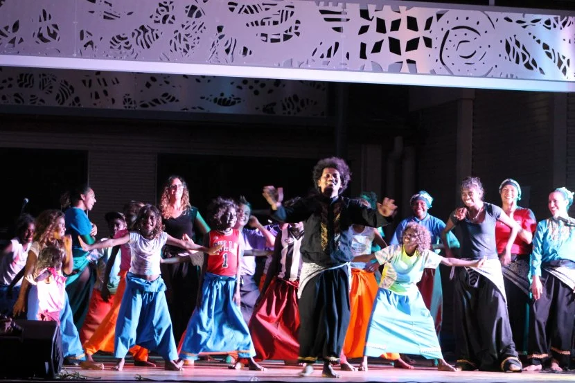 A group of children and adults dancing on stage, holding hands. The stage has decorative cut-out panels and colorful lighting.