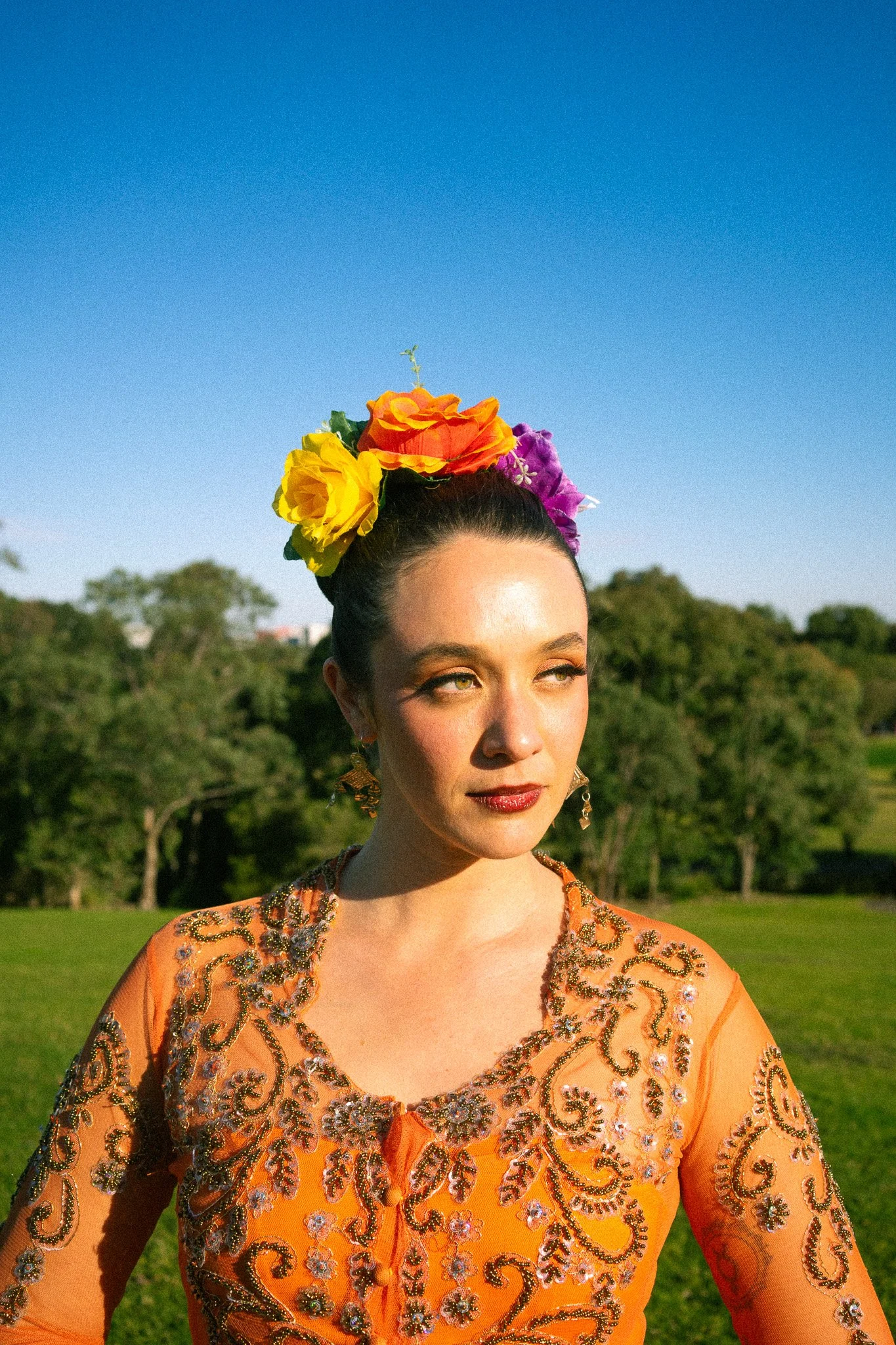 A young woman with dark hair styled in a bun, wearing colorful flowers on her head, gold earrings, and a vibrant orange embroidered dress outdoors on a sunny day.