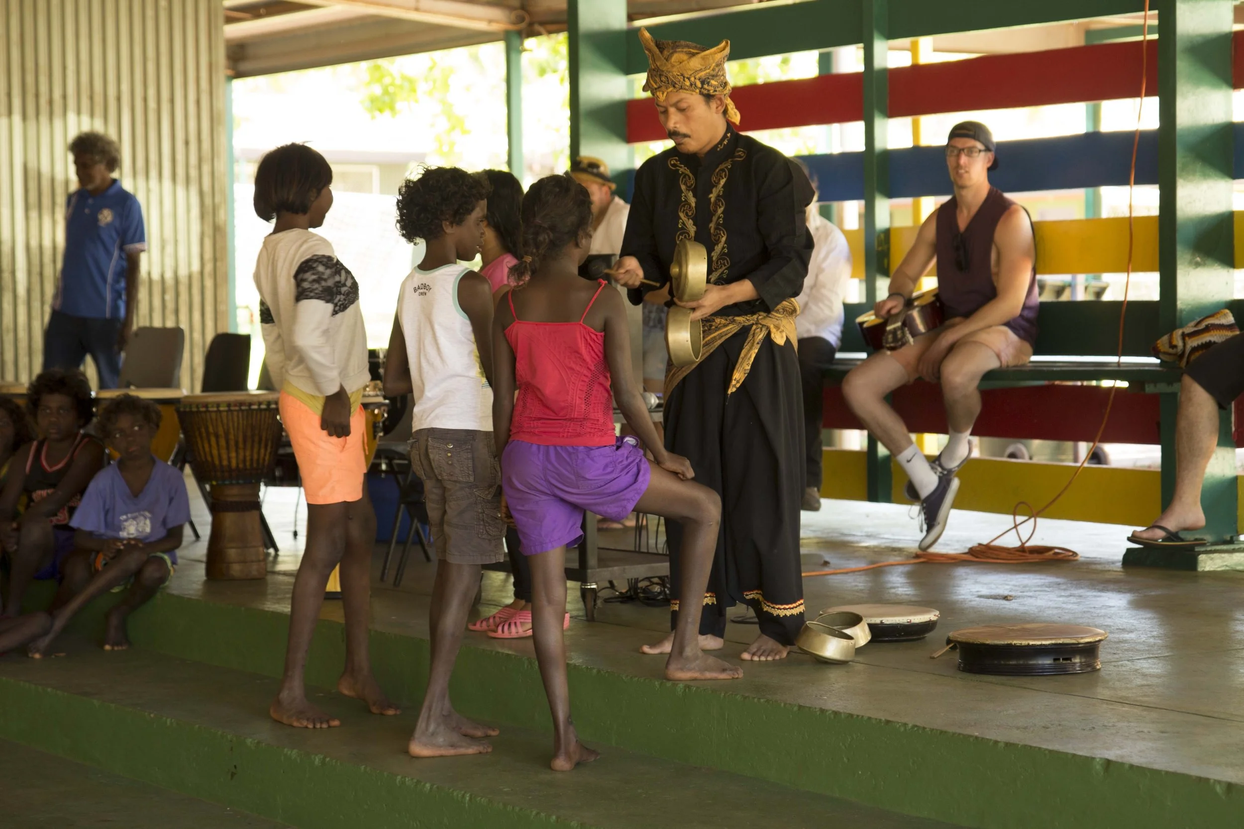 A man dressed in traditional attire leading a cultural performance for children, with seated musicians in the background, on a stage at an indoor venue.
