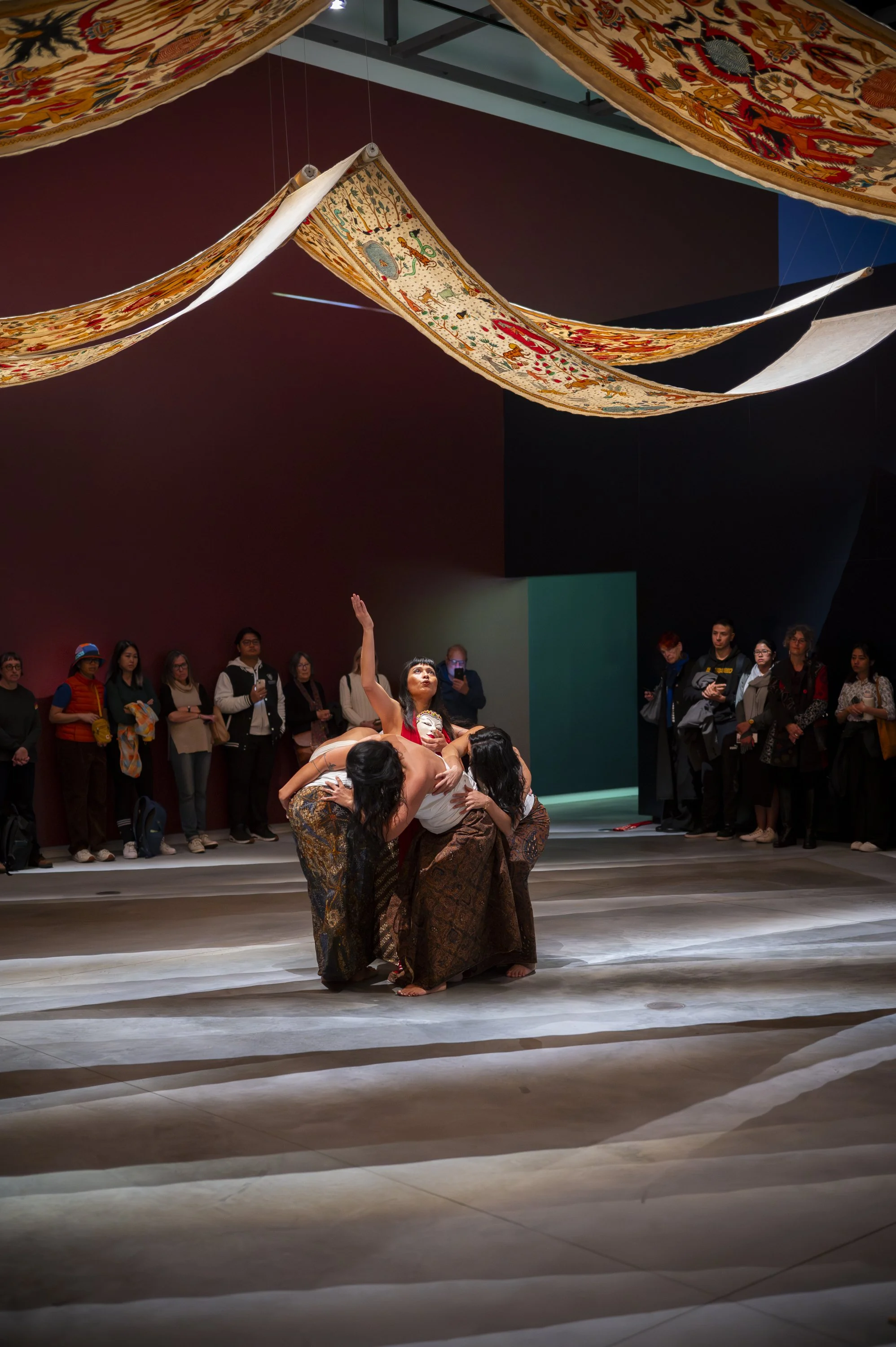 A group of performers is engaged in a dance or theatrical performance on a stage with an audience watching. The stage decor includes hanging ornate fabric banners with colorful patterns. The performers are dressed in traditional costumes, some with brown patterned skirts or pants, and one person with their face painted, possibly as part of a cultural or artistic expression.