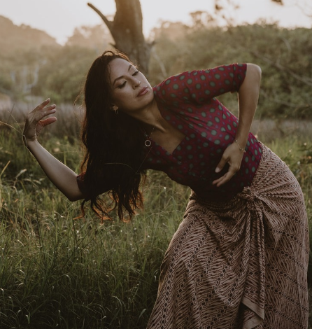 A woman with long dark hair poses outdoors in a grassy field during sunset, wearing a red and green polka dot top and a patterned skirt.