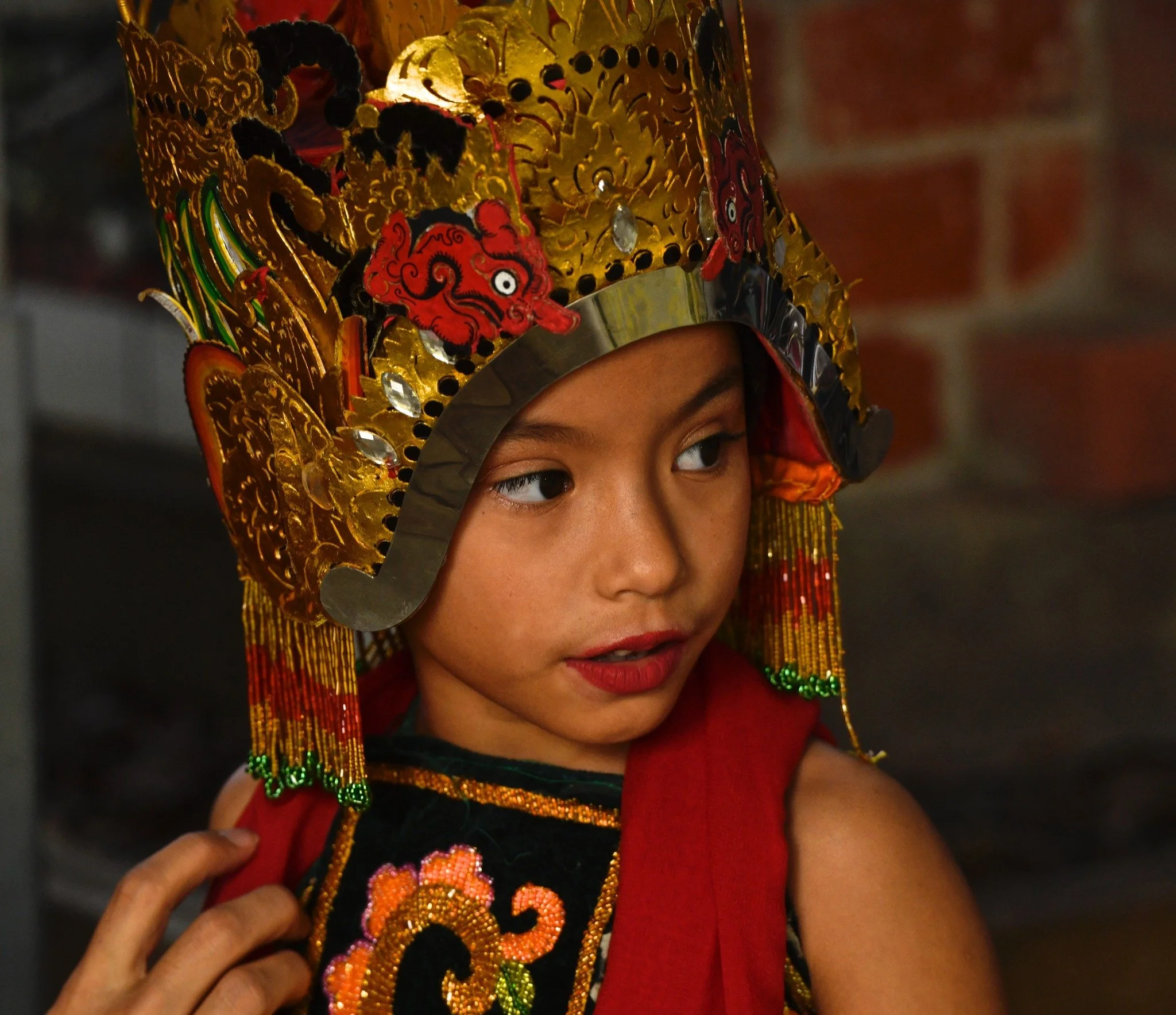 A young girl wearing a traditional colorful headdress with intricate gold, red, and green decorations, and a black embroidered garment with floral designs.
