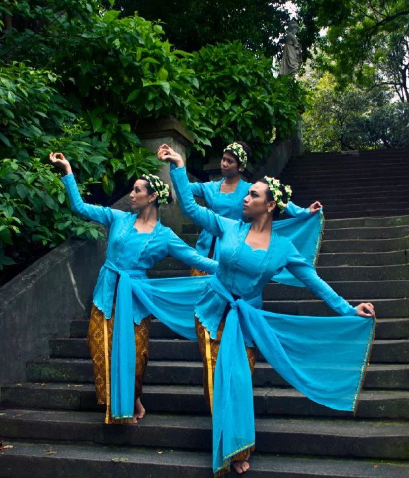 Three women dressed in traditional Indonesian blue and gold costumes perform a dance on outdoor stone stairs surrounded by green foliage.