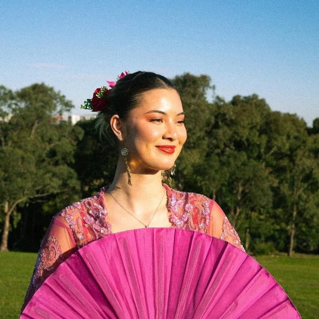 A woman in traditional Mexican dress holding a pink fan in an outdoor park setting with green trees and a clear blue sky.