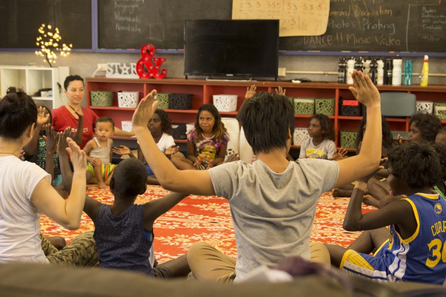 Children and adults sitting cross-legged on a red patterned rug in a circle, raising their hands during an activity in a classroom with shelves, decorations, and a blackboard.