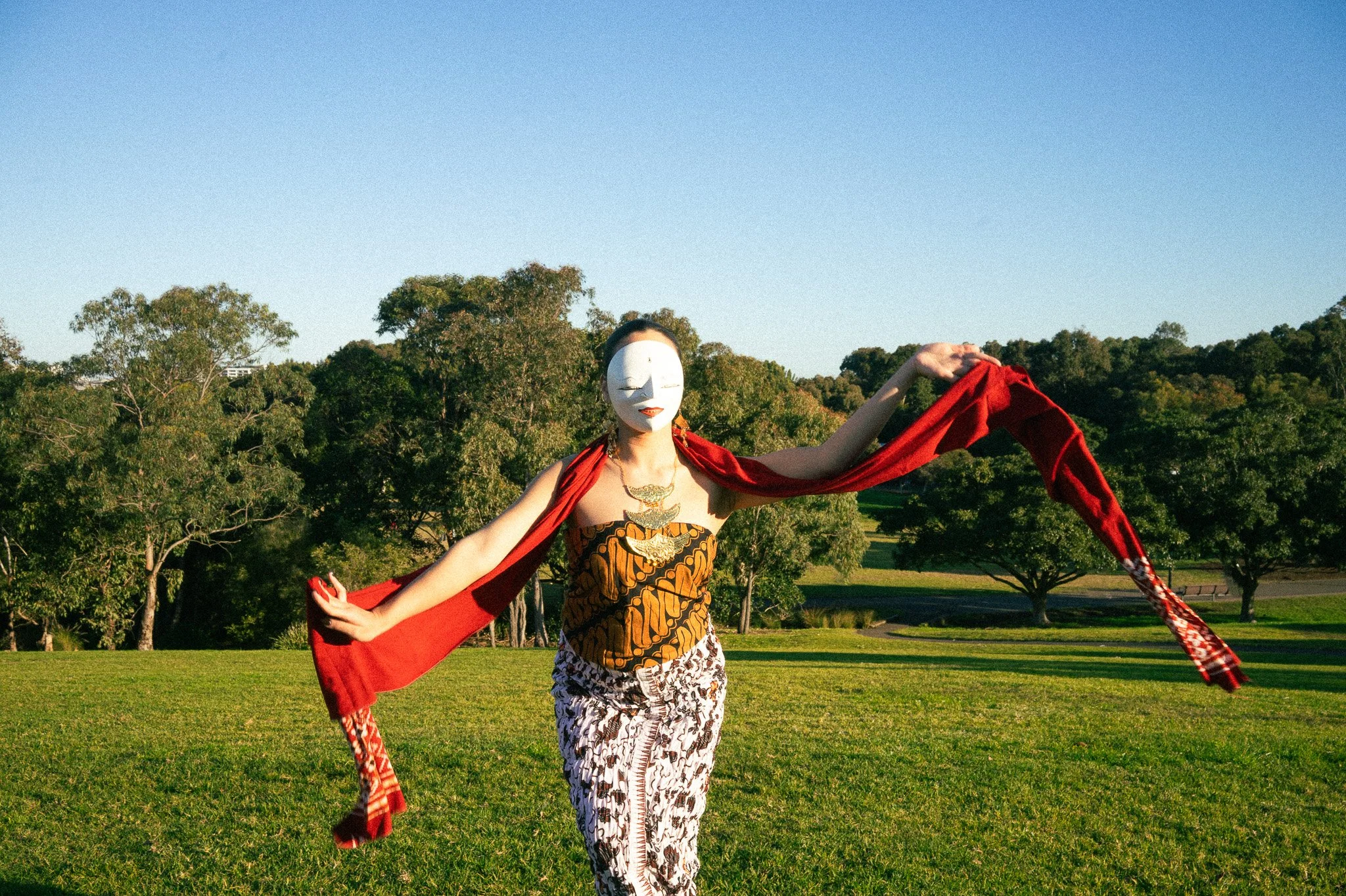 Dancer wearing a white mask, patterned pants, a black and orange top, and a red shawl, standing in a grassy park with trees in the background on a sunny day.
