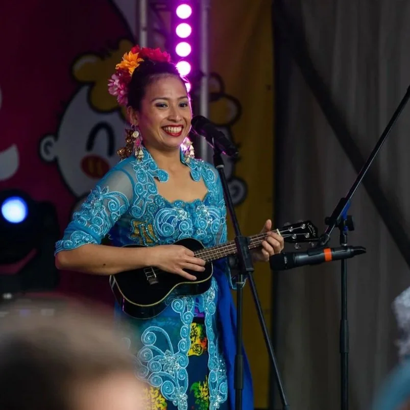 A woman with black hair and a sleeveless patterned top standing outdoors near a body of water, with overhanging tree branches and green foliage in the background.