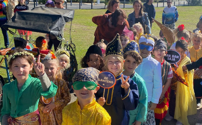 Group of children in colorful costumes at outdoor event with adults, some children wearing accessories like glasses and hats, and holding signs.