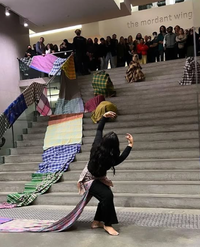 A woman with long black hair performing on a staircase in front of an audience, with colorful fabrics draping the stairs as part of her act.