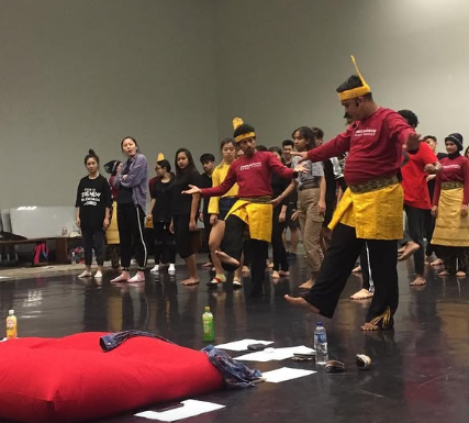 People participating in a traditional dance or performance, some wearing headdresses and costumes, on a stage or rehearsal space.