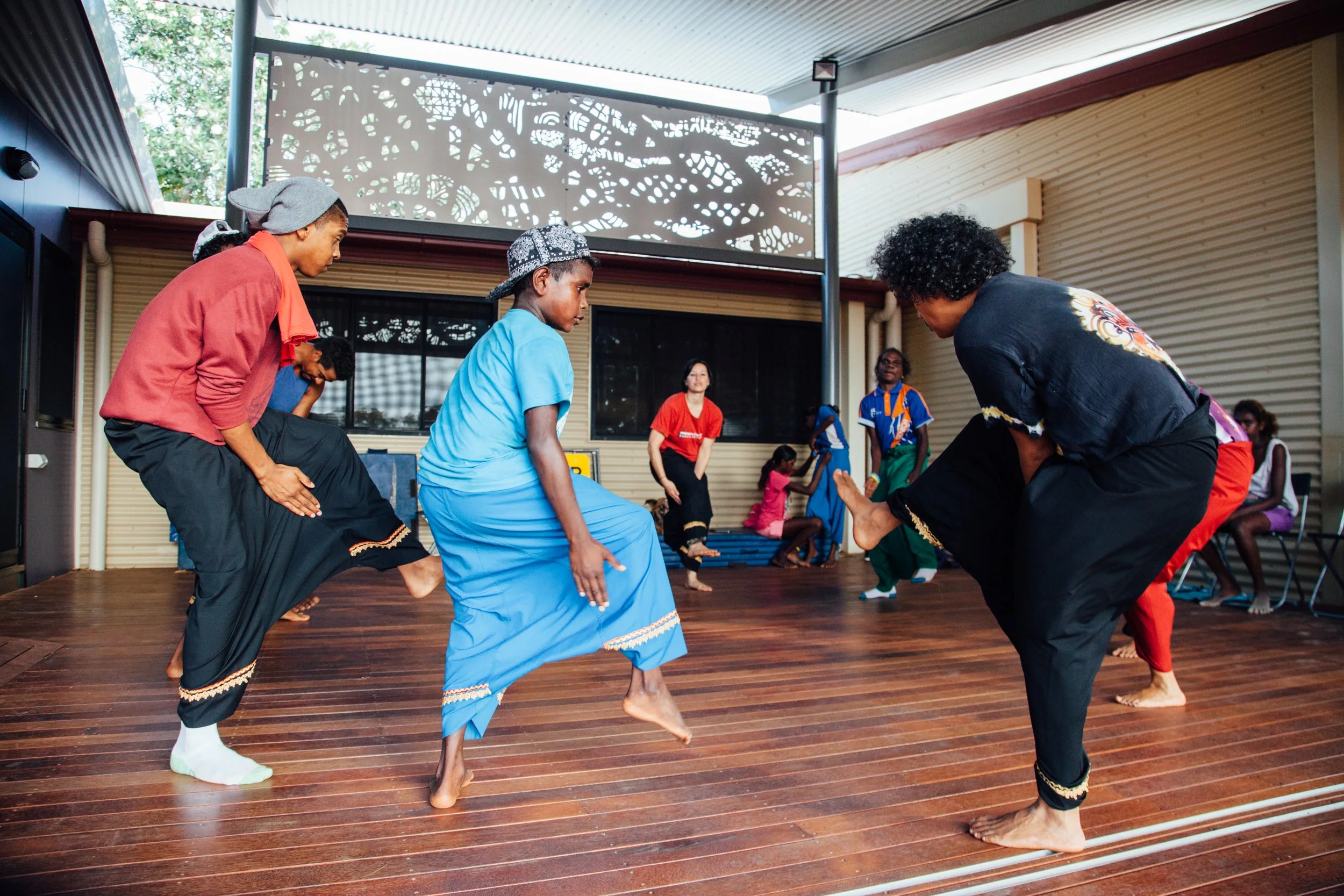 Group of people performing traditional Randai dance on a wooden floor in an indoor space with large windows and decorative panels.