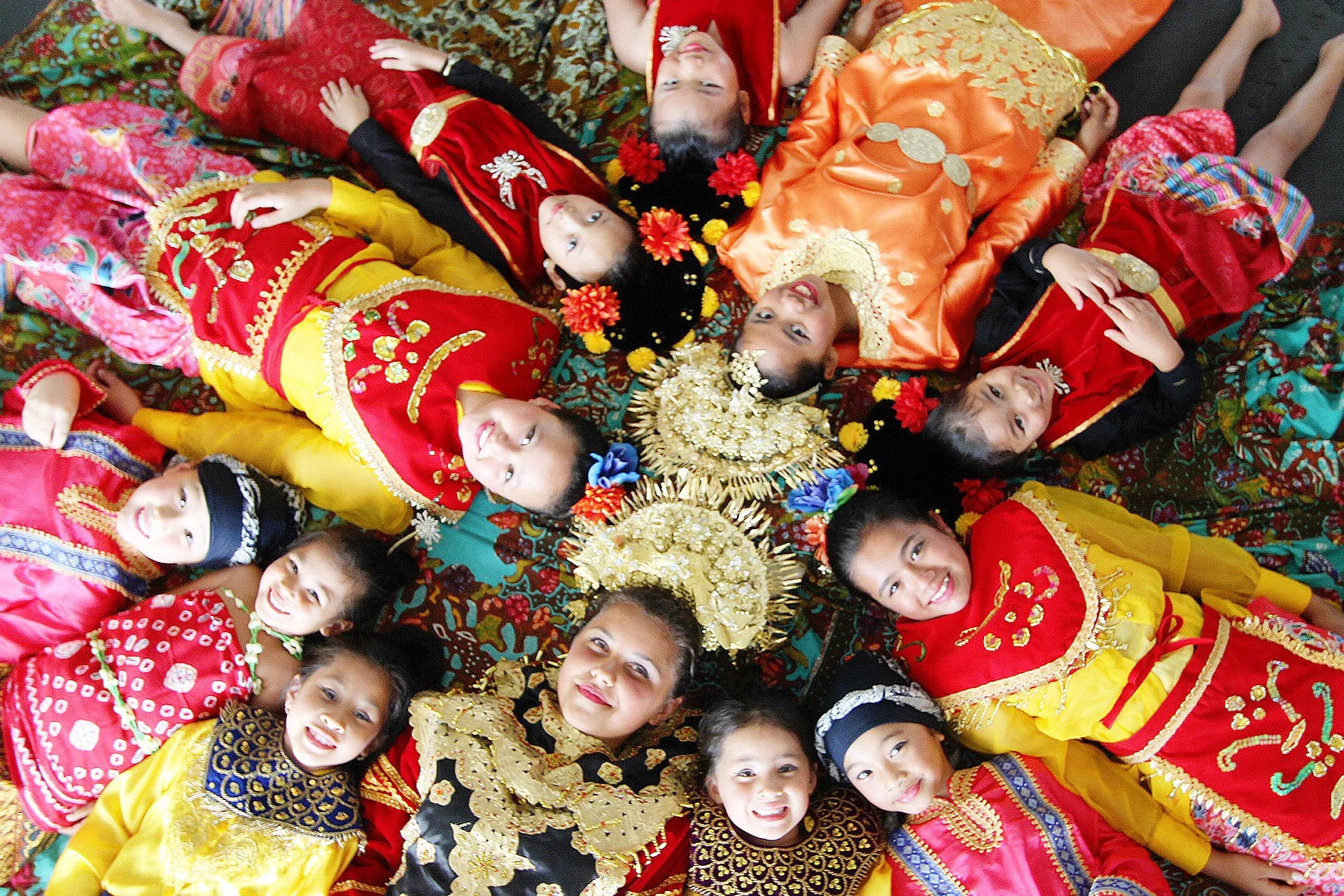 Children dressed in colorful traditional costumes lying in a circle around two women wearing ornate dresses and golden headpieces, with floral decorations in their hair.