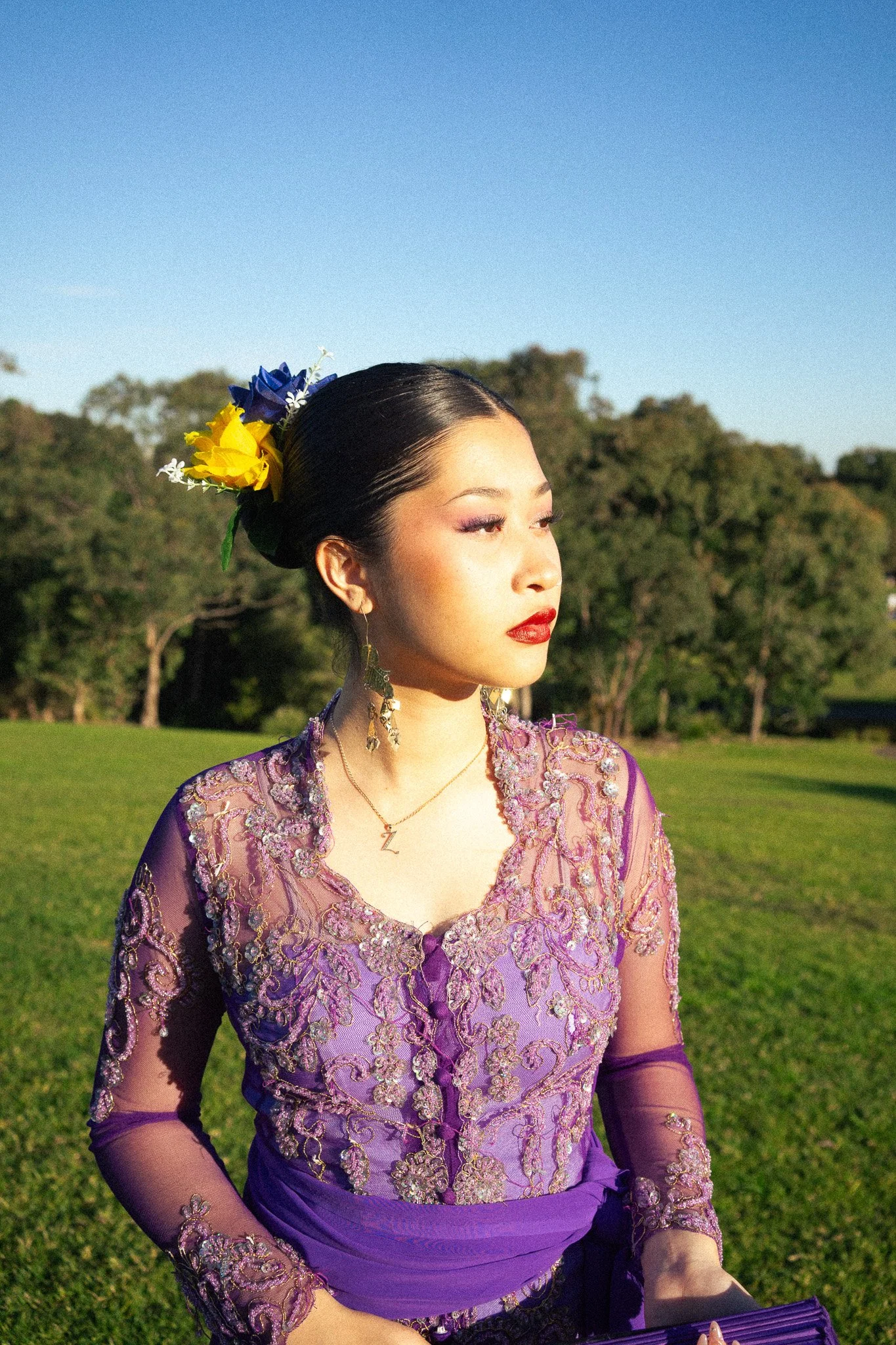 A woman with dark hair styled in an elegant bun, adorned with yellow and blue flowers, wearing a purple lace dress with embroidery, standing outdoors in a park with green grass and trees, during sunset.