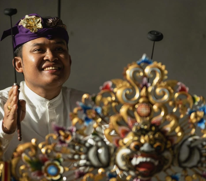 A smiling man wearing traditional Balinese attire, including a purple headscarf and white shirt, behind an ornate, colorful instrument with intricate gold and floral designs.