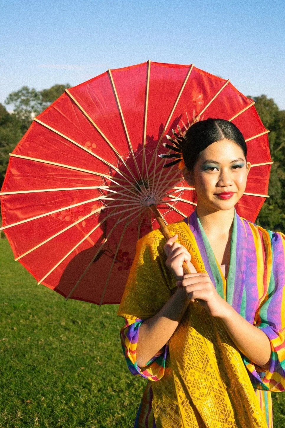 Young woman dressed in traditional Asian attire holding a red paper umbrella outdoors on a sunny day with green trees in the background.