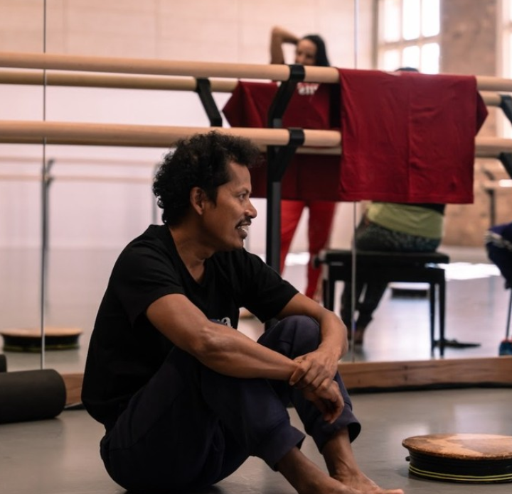 A man sitting on the floor with his knees drawn up in a dance studio, smiling. In the background, a woman in red workout clothes looks into a mirror near ballet barres and chairs.