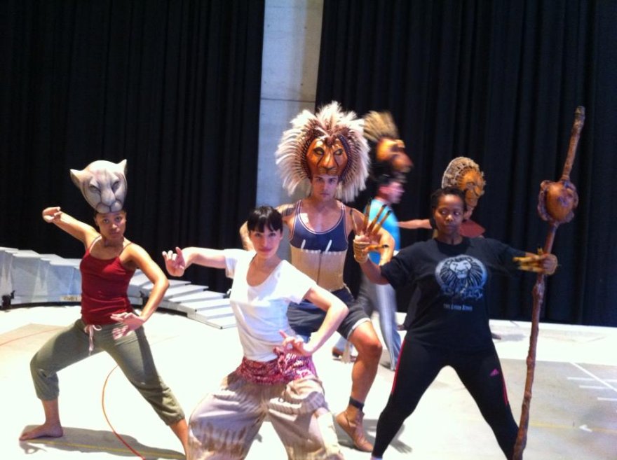 The image shows group of performers in costume on stage, with some wearing lion and animal masks and headdresses, posing in theatrical or dance positions.