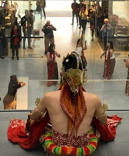 A person dressed in traditional Indian attire with a headpiece and decorated in vibrant colors, sitting on a raised platform in front of a large mirror in an indoor mall or gallery. Reflecting in the mirror, multiple onlookers, some taking photos and others observing, can be seen.