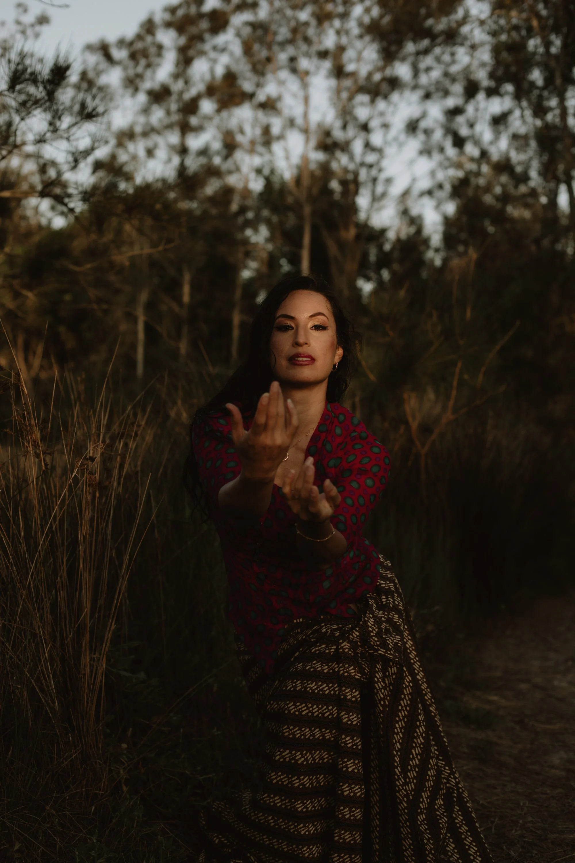 A woman with dark hair and makeup, wearing a red and green polka dot blouse and a patterned skirt, standing outdoors in front of trees during sunset, gesturing with her hands.